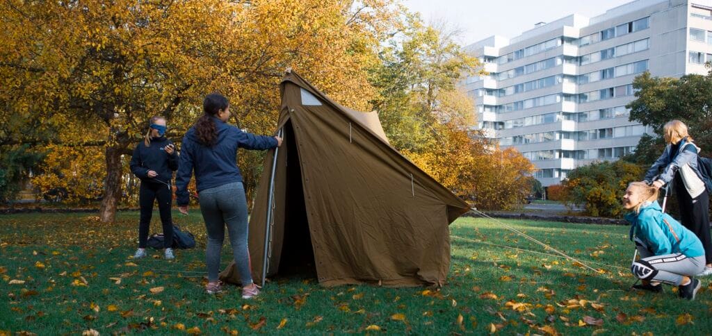 Neljä nuorta pystyttämässä telttaa nurmikolla. Taustalla syksyisiä lehtipuita ja kerrostalo.